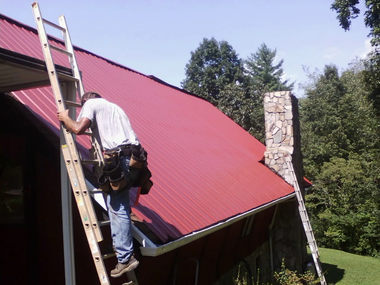 Roofer on a ladder beside a red metal roof and stone chimney — Soffit & Fascia Repair by Roofing Brothers in Lynn Center